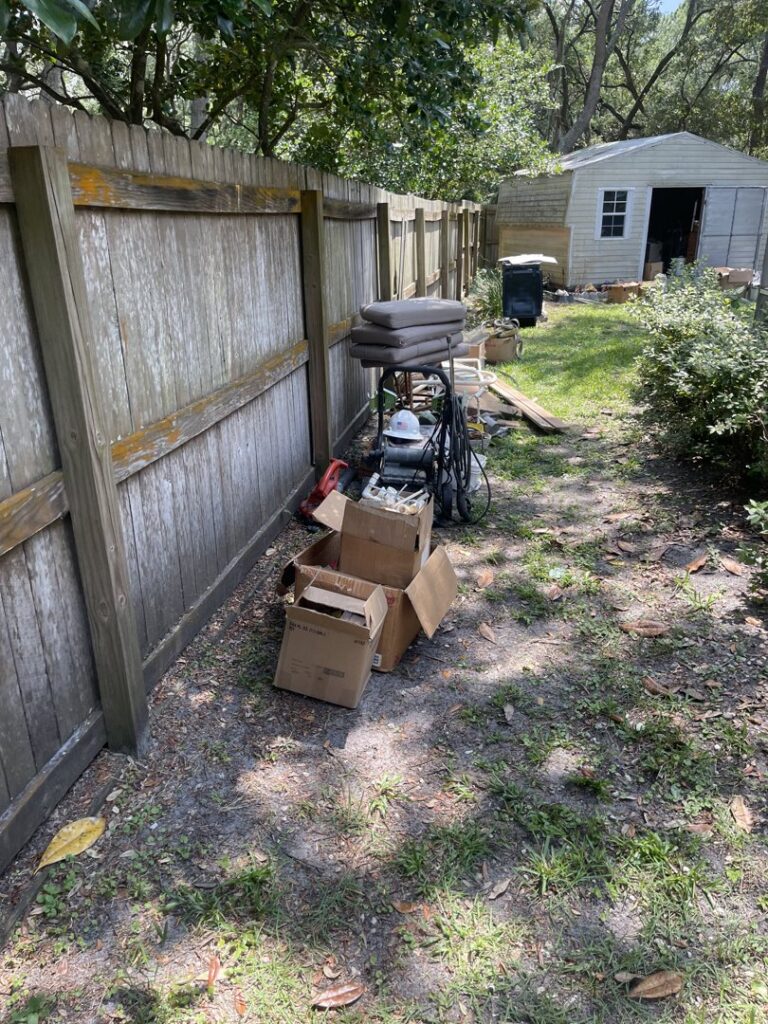 Boxes and various items piled up against a wooden fence in a backyard, ready for junk removal by Jacksonville Hauling & Junk Removal in Jacksonville, FL.