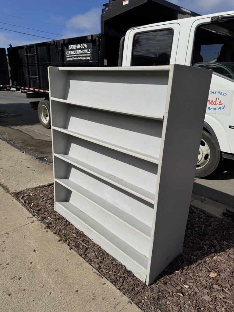 A bookshelf placed on the sidewalk next to a Fred's Junk Removal truck in Chula Vista, CA.