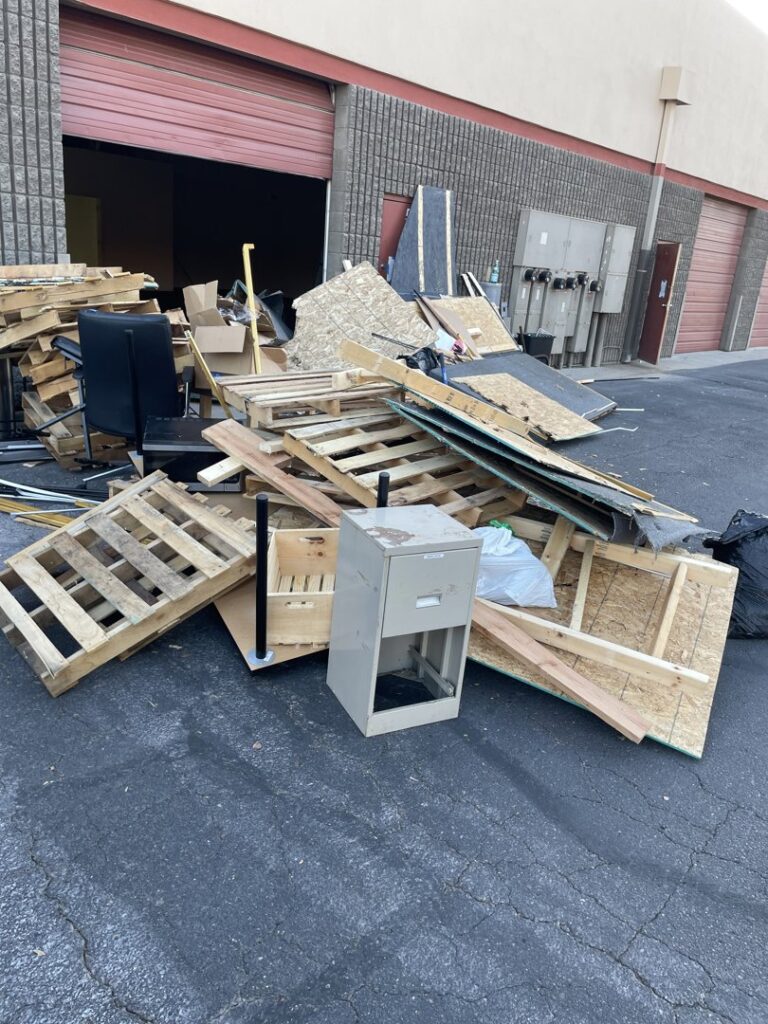 A large pile of wooden pallets, plywood, and a filing cabinet outside a commercial building, ready for junk removal by Cee J Junk Removal LLC in Phoenix, AZ.