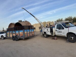 A crane lifting a large metal tank onto a North Valley Junk Removal branded trailer during a heavy junk removal job in Phoenix, AZ.