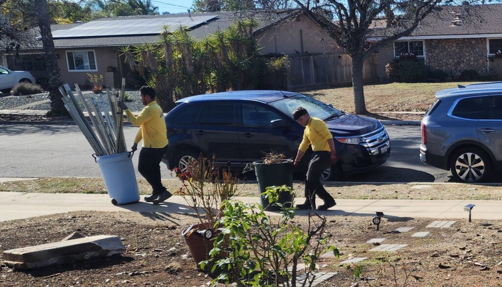 Crisan Junk Removal workers hauling away metal poles and yard debris in trash cans in San Diego, CA.