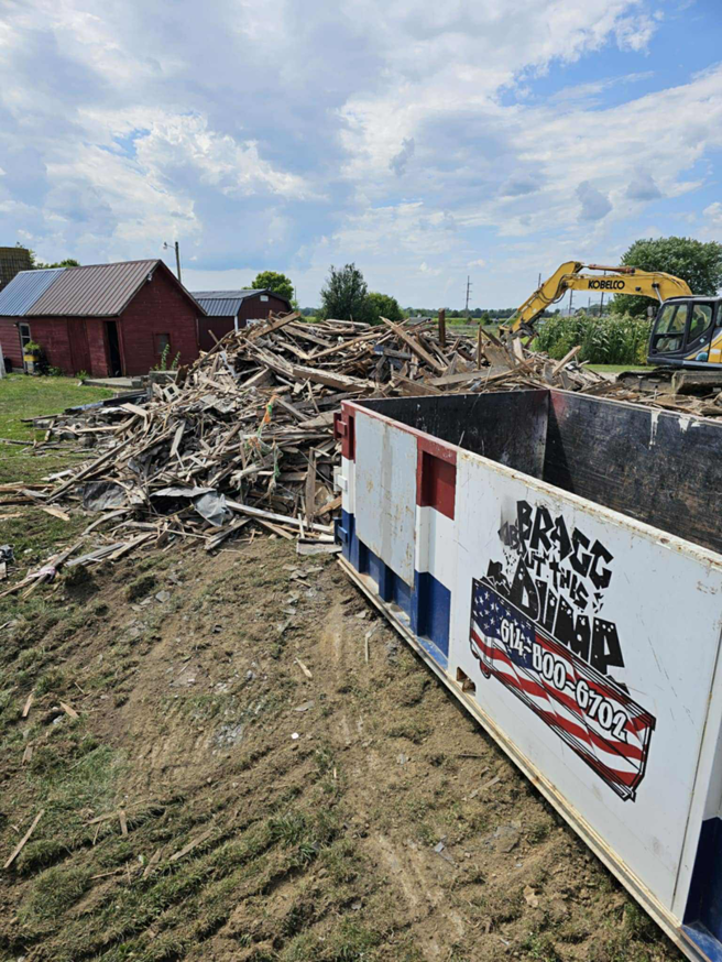 A large pile of demolition debris being loaded into a branded dumpster by Bragg About This Dump in Columbus, OH, with an excavator nearby.