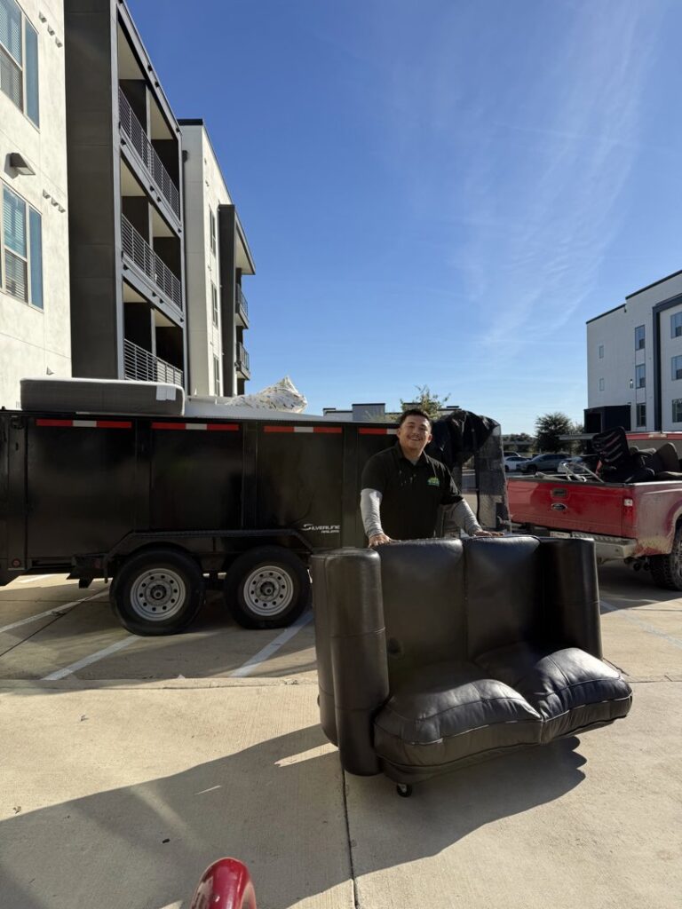 A JunkGuys Austin Junk Removal employee is moving a sofa, with a loaded junk removal trailer visible in the background in Austin, TX.