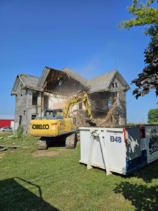 An excavator demolishing an old house, with a branded dumpster from Bragg About This Dump positioned nearby for debris removal in Columbus, OH.