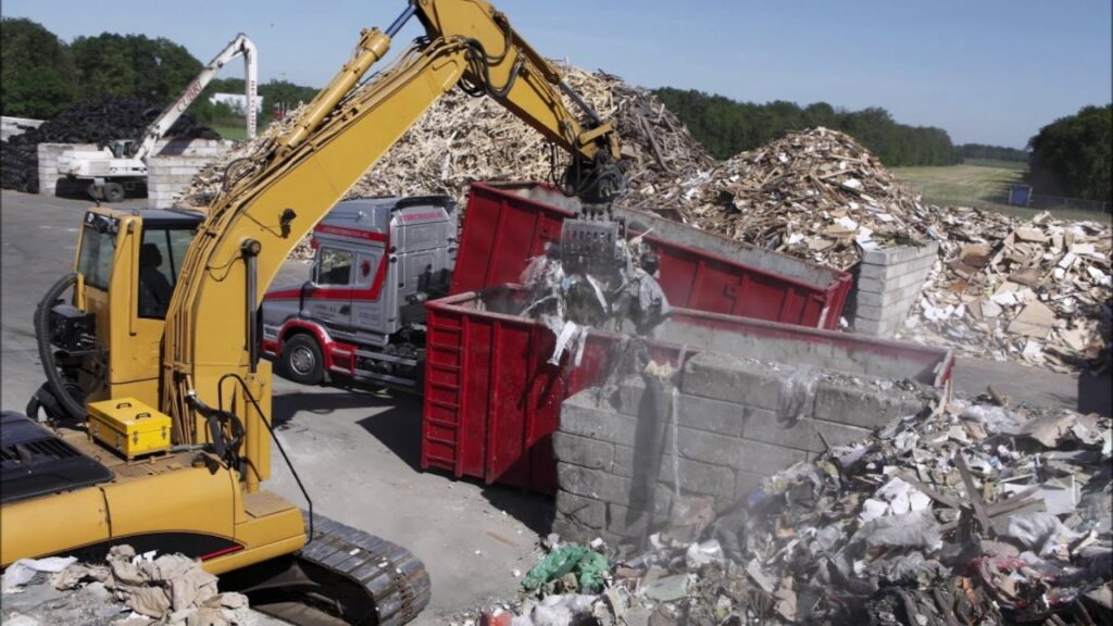 An excavator loading a large pile of mixed waste into a red dumpster at a facility, demonstrating junk removal services by Epic Junk Removal SD in San Diego, CA.