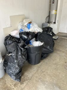 Black trash bags and a bin filled with various junk in a garage, ready for removal by Texas Junk Masters in Fort Worth, TX.