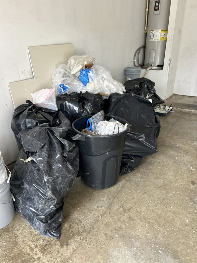 Black trash bags and a bin filled with various junk in a garage, ready for removal by Texas Junk Masters in Fort Worth, TX.