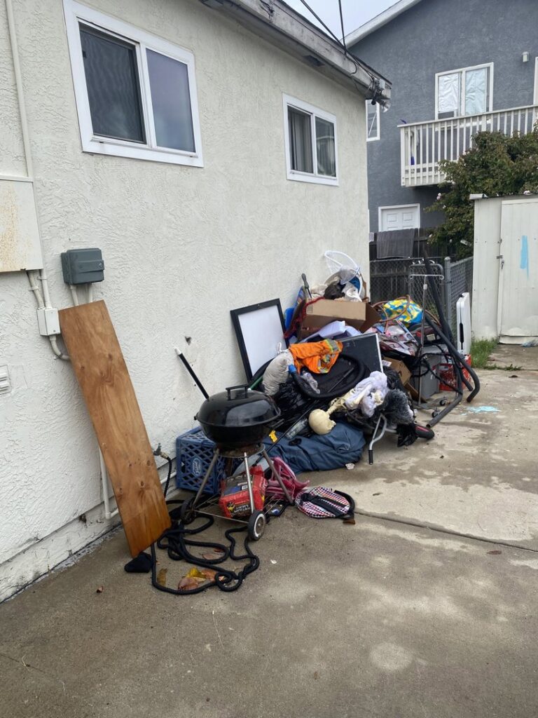 A pile of miscellaneous household junk, including a grill and bicycle, next to a house for Crisan Junk Removal in San Diego, CA.