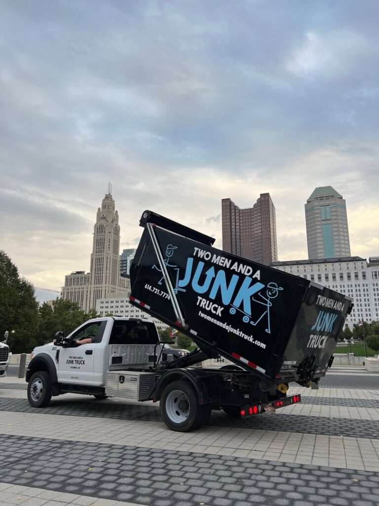 A branded junk removal truck from Two Men and a Junk Truck with its dump bed raised, against a city skyline in Columbus, OH.