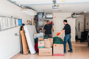 Two JunkStart crew members preparing to remove a large pile of clutter, including a mattress and boxes, from a garage in San Antonio, TX.