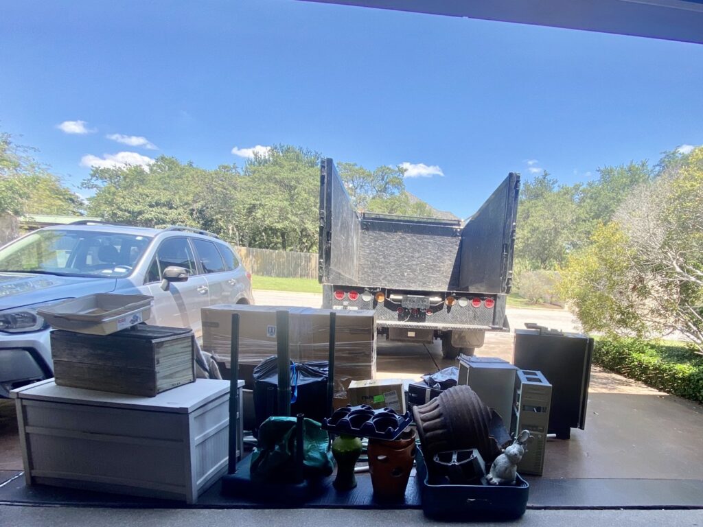 A large pile of junk, including a TV, bicycles, and trash bags, on a grassy area next to a sidewalk in Austin, TX.