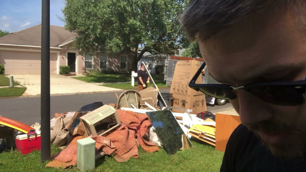 A large pile of household junk and debris on a residential lawn, with a worker in the background, indicating a junk removal job by Jacksonville Hauling & Junk Removal in Jacksonville, FL.