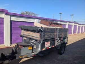 A Herrera Hauling & Junk Removal trailer loaded with furniture and junk, parked in front of storage units in Fort Worth, TX.