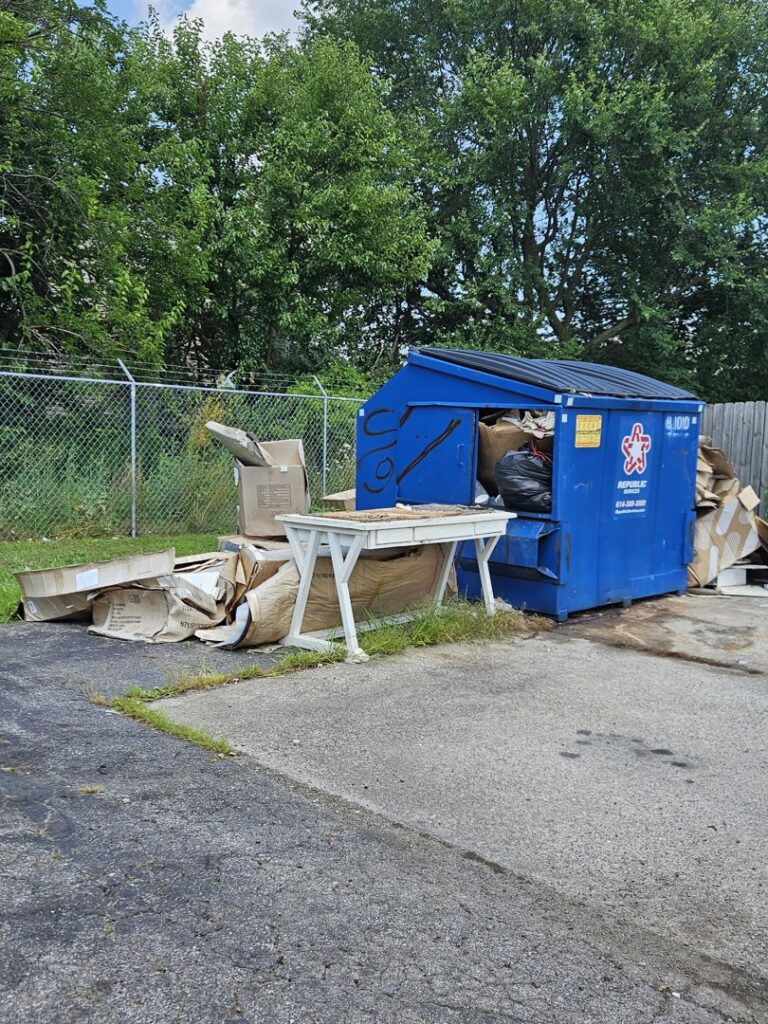An overflowing blue commercial dumpster surrounded by cardboard boxes and debris, indicating a large junk removal job by Purefoy's Professionals in Columbus, OH.