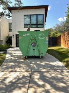 A green 1-800-JUNKPRO dumpster rental, partially filled with junk, in a residential driveway in Austin, TX.