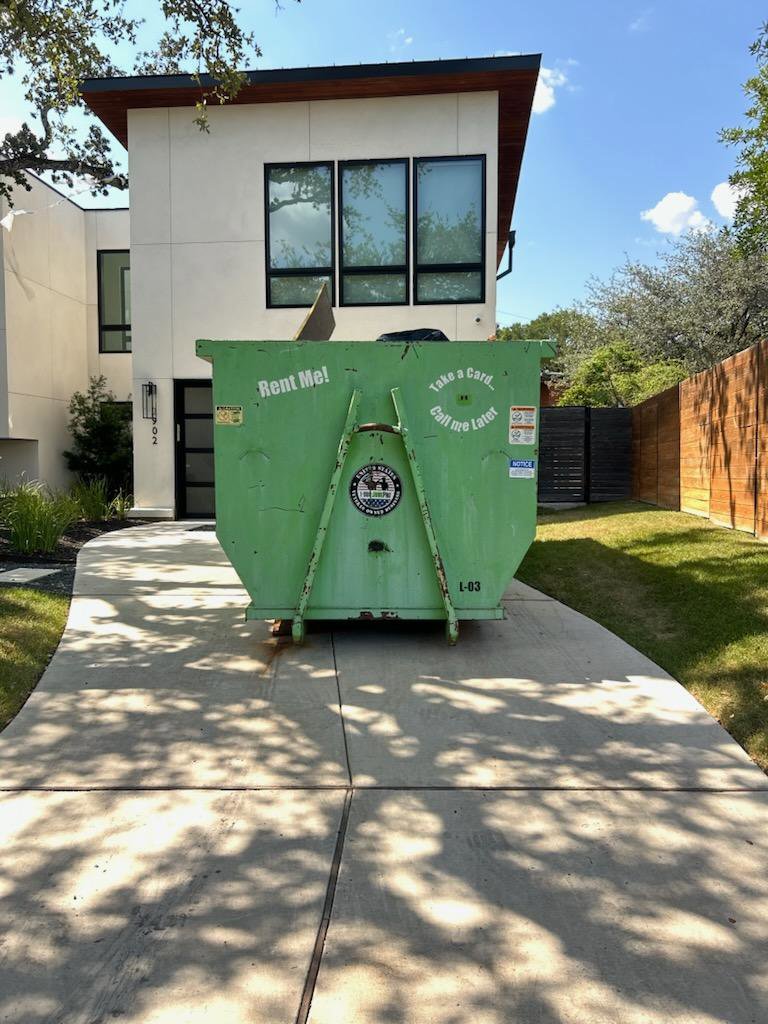 A green 1-800-JUNKPRO dumpster rental, partially filled with junk, in a residential driveway in Austin, TX.