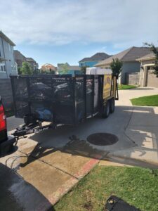 A large pile of various junk items on a sidewalk next to a trailer from All Needz Junk Removal & Hauling Service in Dallas, TX, ready for pickup.