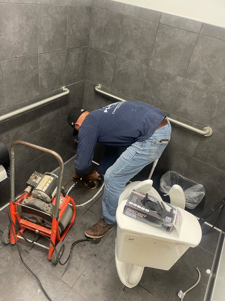 A plumber from Plumbing Services Inc. performing toilet repair or installation with a drain snake machine nearby in San Diego, CA.