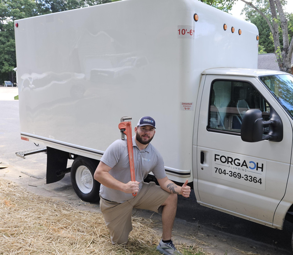 A plumber from Forgach Plumbing LLC in Charlotte, NC, kneeling with a large pipe wrench next to a branded service truck.