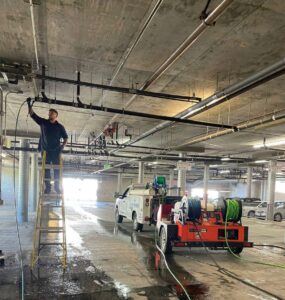 A plumber from Plumbing Services Inc. working on overhead pipes in a commercial building, with hydro-jetting equipment on site in San Diego, CA.