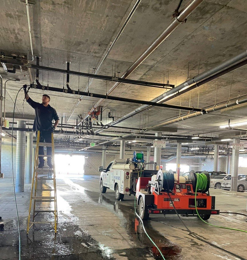 A plumber from Plumbing Services Inc. working on overhead pipes in a commercial building, with hydro-jetting equipment on site in San Diego, CA.