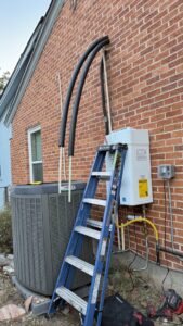An outdoor tankless water heater installation site with a ladder and AC unit by Economy Plumbing Services in Austin, TX