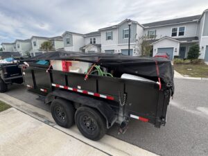 A tarped dump trailer filled with various items of junk, parked on a residential street for Jax Junkies Removal LLC in Jacksonville, FL