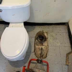 A toilet removed, showing the drain flange and a drain cleaning auger, indicating a plumbing service by Tribeca Plumbing in Dallas, TX.