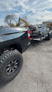 A Faithful Hands Junk Removal truck and trailer in San Antonio, TX, hauling a load of scrap metal and debris, with an excavator in the background.