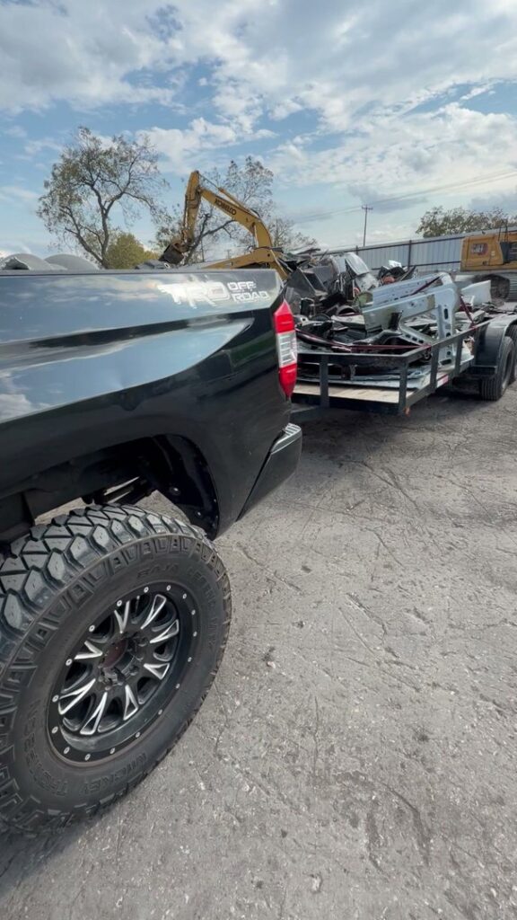 A Faithful Hands Junk Removal truck and trailer in San Antonio, TX, hauling a load of scrap metal and debris, with an excavator in the background.