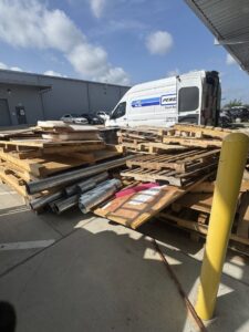 A large stack of wooden pallets and metal pipes next to a white commercial van, ready for removal by HustleTown Junk Removal in Houston, TX.
