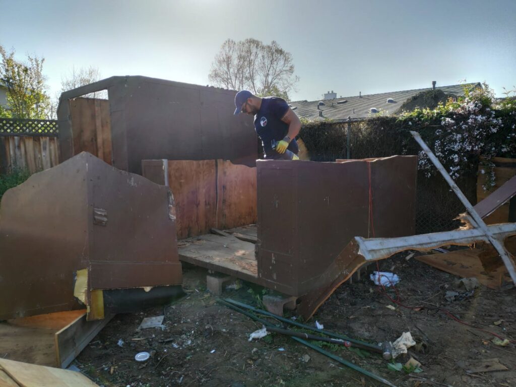A worker dismantling an old wooden shed in a backyard for Strong Hands Hauling & Dumpster Rental in San Jose, CA