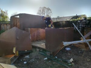 A worker dismantling an old wooden shed in a backyard for Strong Hands Hauling & Dumpster Rental in San Jose, CA