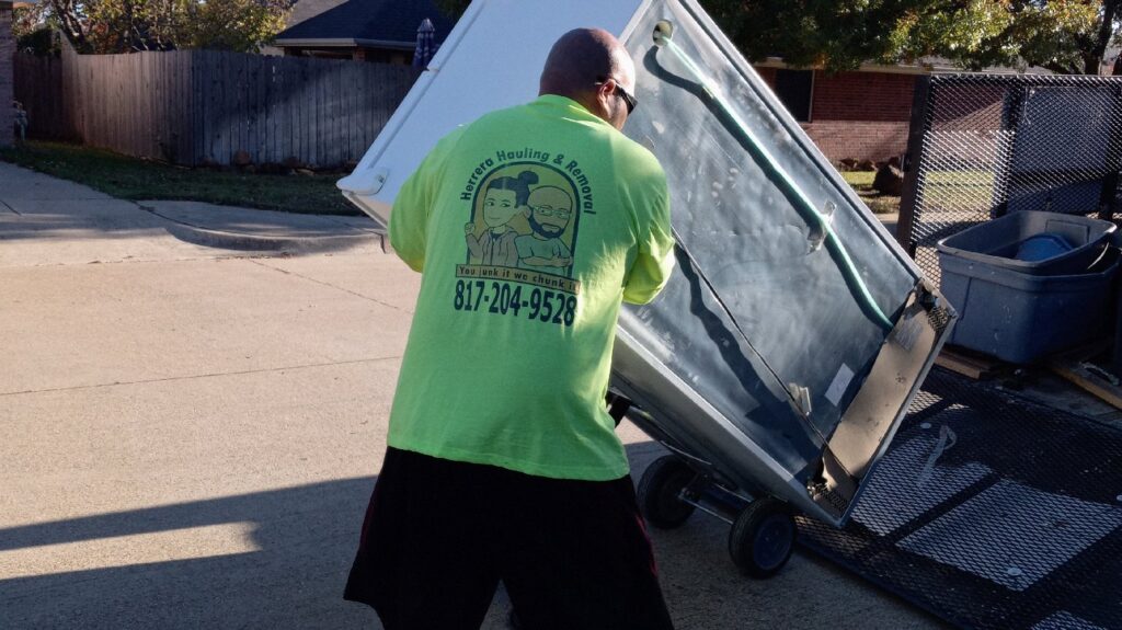 A worker from Herrera Hauling & Junk Removal loading a large refrigerator onto a trailer in Fort Worth, TX.