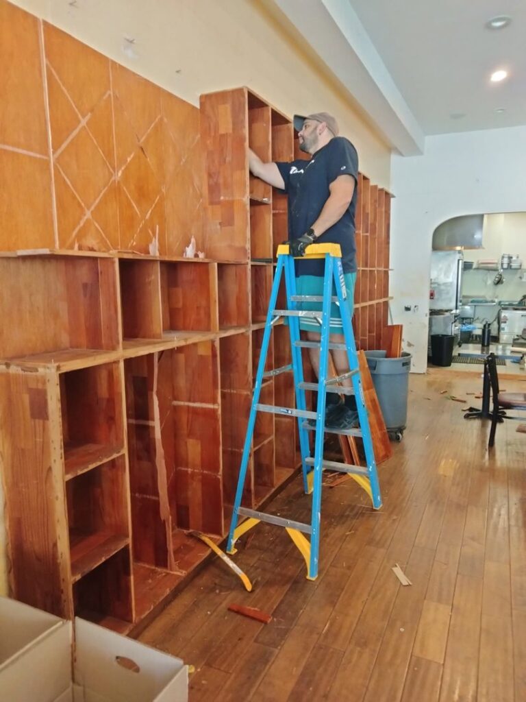 A worker removing wooden shelving from a wall for Strong Hands Hauling & Dumpster Rental in San Jose, CA