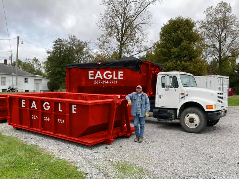 An Eagle Dumpster Rental worker standing next to a truck and two red dumpsters, one covered, in Philadelphia, PA.