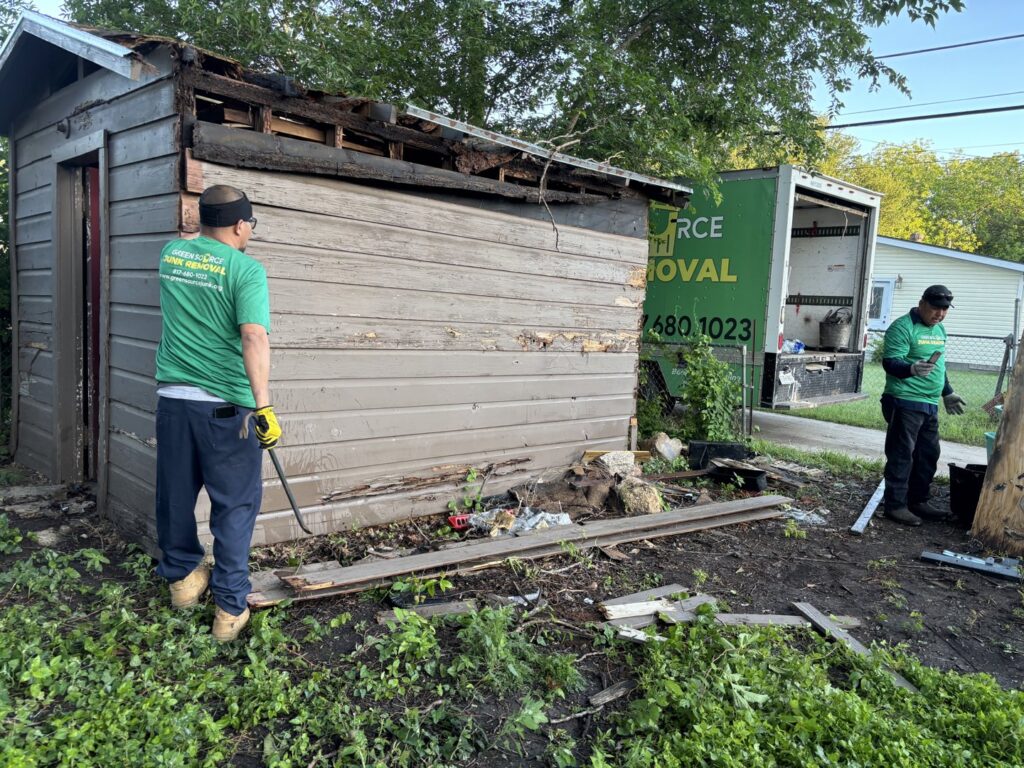 Two Green Source Junk Removal workers demolishing an old shed with their truck visible in the background in Fort Worth, TX.