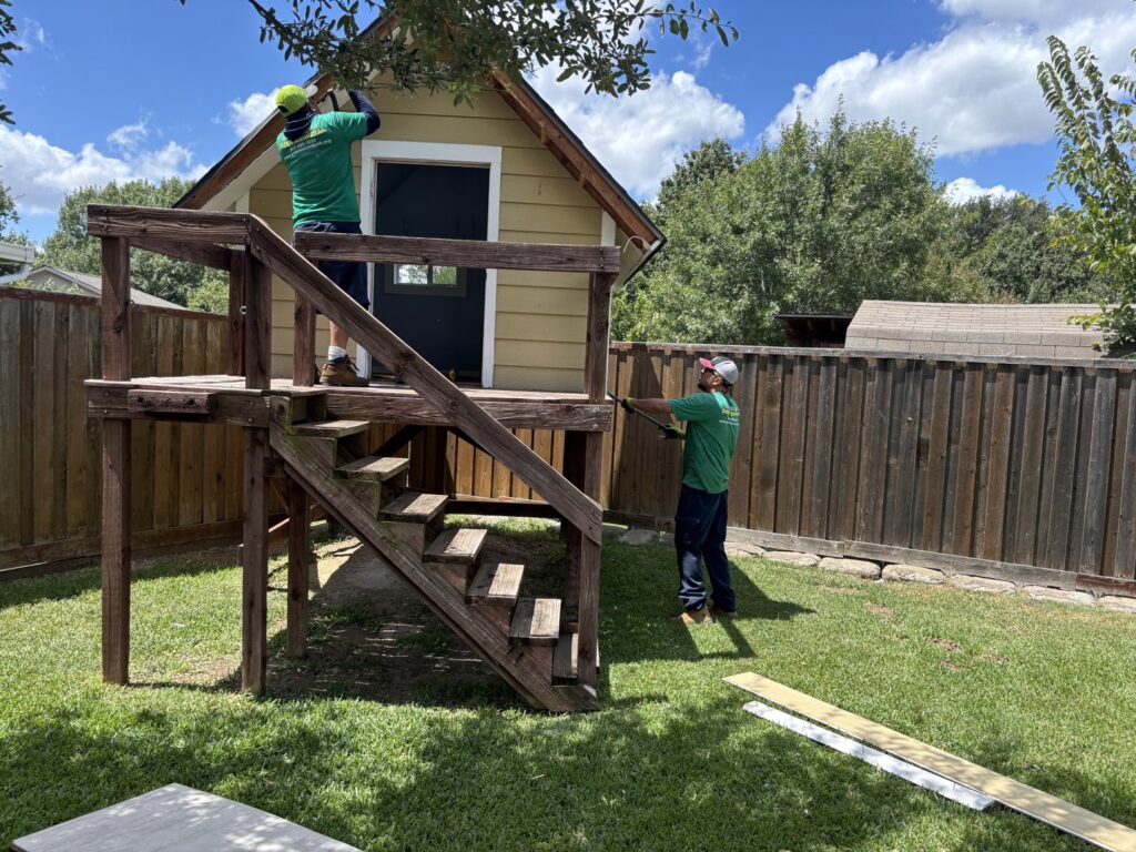 Two Green Source Junk Removal workers dismantling a wooden playhouse structure in a backyard in Fort Worth, TX.