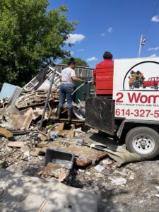 Two workers from 2 Women with a Pickup Truck and Trailer Too LLC loading a large pile of debris into a dump trailer during a junk removal job in Columbus, OH.