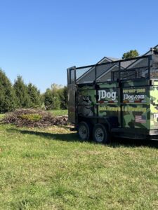 A JDog Junk Removal & Hauling trailer parked next to a large pile of tree branches and yard waste in Columbus, OH.